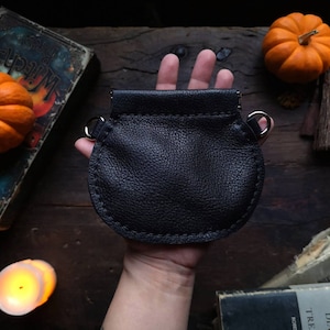 May include: A black leather pouch with a silver clasp and a drawstring closure. The pouch is held in a hand against a dark wooden background.