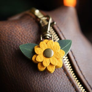 May include: Close-up of a brown leather bag with a gold zipper. A yellow leather flower charm with green leaves and a brass center is attached to the zipper pull. The bag has a textured surface.