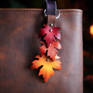 Peut inclure: Un porte-clés en cuir avec trois breloques en forme de feuilles d'érable rouges, oranges et jaunes. Les feuilles sont attachées à une lanière en cuir marron et à un anneau argenté. Le porte-clés est suspendu à un sac en cuir marron.