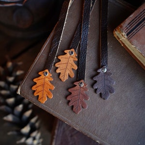 May include: Four leather bookmarks with oak leaf designs in brown, tan, and dark brown. The bookmarks are on a brown leather-bound book.