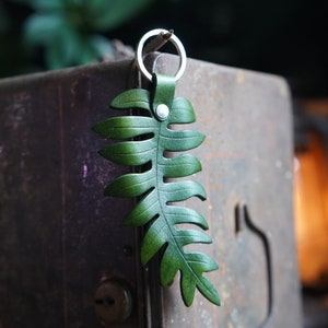 A green leather fern keychain is balanced on a dried fern frond on the edge of a rusted metal box. There is a lantern glowing in the background.