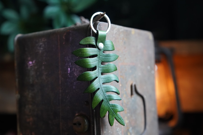 A green leather fern keychain is balanced on a dried fern frond on the edge of a rusted metal box. There is a lantern glowing in the background.