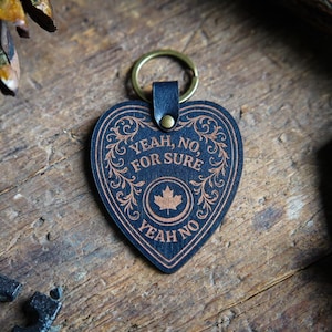 May include: A heart-shaped keyring with the text "YEAH, NO, FOR SURE YEAH NO" and a maple leaf symbol. The keyring is black with a brown decorative border and text. It has a leather strap and a gold-coloured ring.