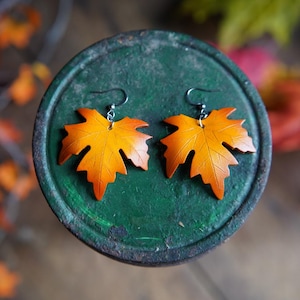 May include: A pair of maple leaf earrings. The earrings are made of orange and yellow material, with a gradient effect. The earrings are hanging from silver-colored hooks. The earrings are resting on a green surface.