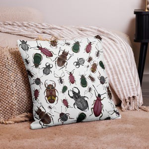 May include: White square throw pillow with a repeating pattern of various colorful beetles and insects. The insects are in shades of brown, green, red, and gray. The pillow is on a bed with a beige blanket.
