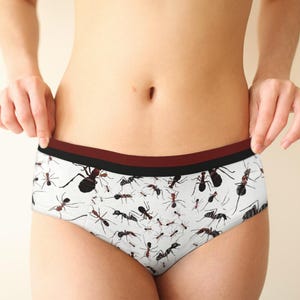 May include: White underwear with a repeating pattern of black and red ants. The waistband is a combination of burgundy and black. The underwear is made of a soft, comfortable material.