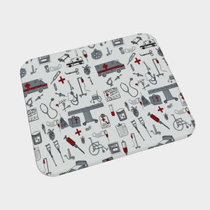 May include: A white rectangular mat with a repeating pattern of medical equipment in gray, red, and black. The pattern includes ambulances, stethoscopes, syringes, and medical crosses. The mat has rounded corners.