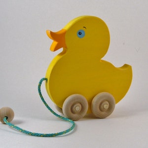 May include: A yellow wooden pull toy shaped like a duck with blue eyes and an orange beak. The duck has two wooden wheels and a rope attached to a wooden ball.