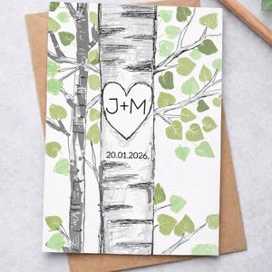 May include: A hand-drawn wedding invitation card featuring a birch tree design with a heart-shaped carving containing the initials "J+M". The date "20.01.2026" is below the heart. The card is on a brown envelope.
