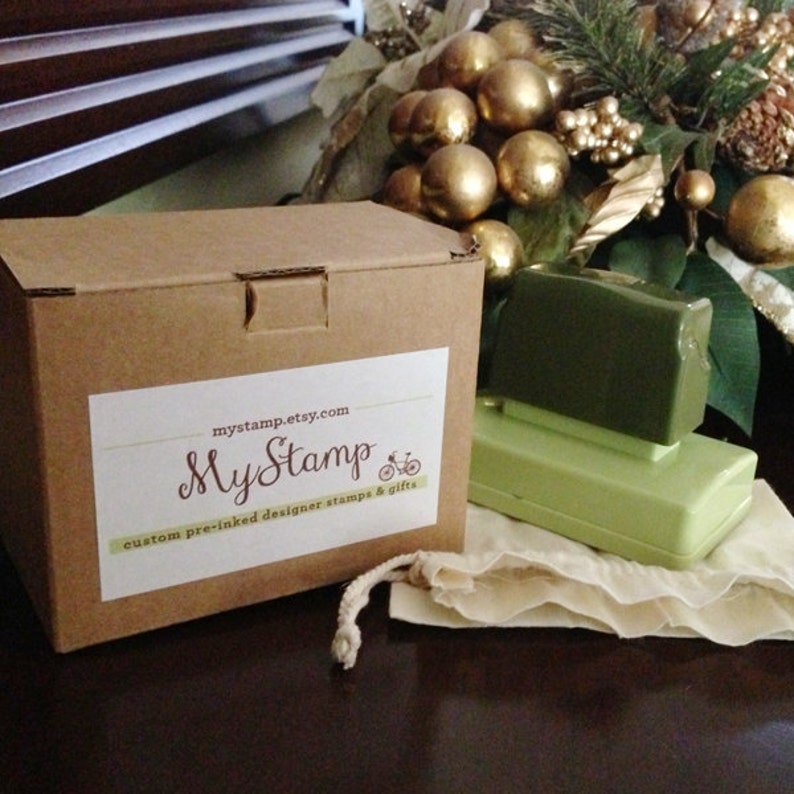 以下が含まれることがあります： 「MyStamp」と「custom pre-inked designer stamps & gifts」と書かれた白いラベルが付いた茶色の段ボール箱。箱の隣には2つの緑色のスタンプと小さなベージュのバッグがあります。背景には金の装飾品と緑があります。