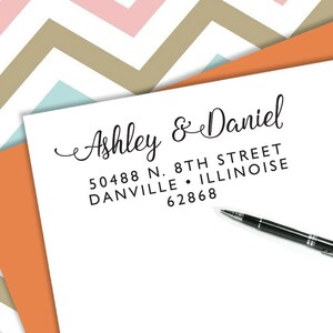 以下が含まれることがあります： 「Ashley & Daniel」の名前と住所がエレガントな筆記体で書かれた白い紙。黒いペンが紙の上に置かれています。背景には、ピンク、タン、ブルーのシェブロン柄があります。
