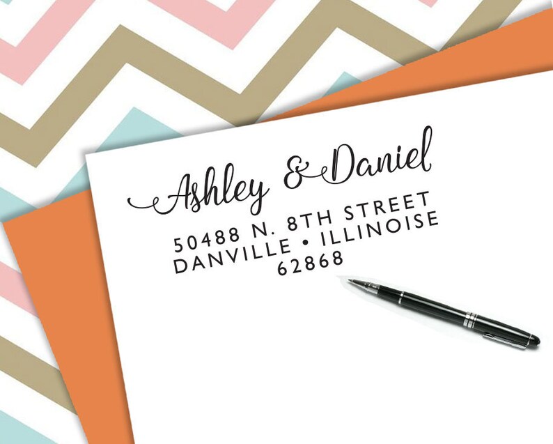 以下が含まれることがあります： 「Ashley & Daniel」の名前と住所がエレガントな筆記体で書かれた白い紙。黒いペンが紙の上に置かれています。背景には、ピンク、タン、ブルーのシェブロン柄があります。