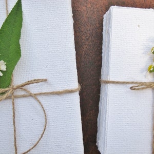 May include: Two stacks of white handmade paper, tied with twine and decorated with small white flowers and green leaves.