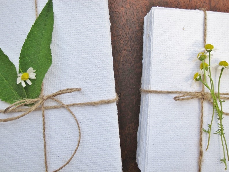 May include: Two stacks of white handmade paper, tied with twine and decorated with small white flowers and green leaves.