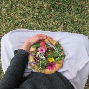 May include: A wooden plate with a variety of flowers and a pair of scissors. The plate is held by a person wearing a white shirt and black pants.