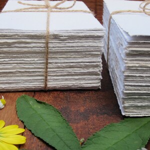 May include: Two stacks of handmade paper, each tied with twine, are arranged on a wooden surface. The paper is white and has a slightly rough texture. There are green leaves and yellow flowers in the foreground.