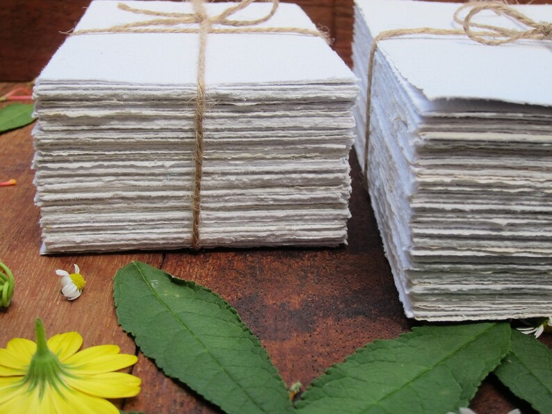 May include: Two stacks of handmade paper, each tied with twine, are arranged on a wooden surface. The paper is white and has a slightly rough texture. There are green leaves and yellow flowers in the foreground.