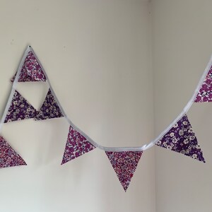May include: A purple and white floral patterned bunting with white string. The bunting is hanging on a white wall.