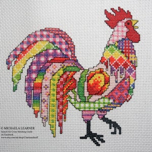 May include: A colorful cross-stitch pattern of a rooster with a pink body, red comb, and a multi-colored tail. The rooster is standing on a white background.