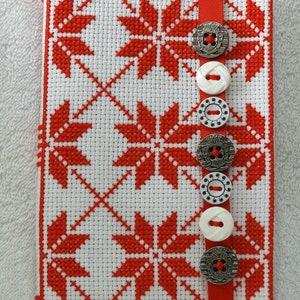 May include: A white and red cross-stitch fabric cover with a red ribbon and silver and white buttons.