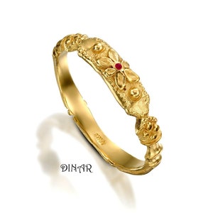 May include: A gold ring featuring a textured band with a central floral motif accented by a small red gemstone and two spherical gold beads. The band has a matte finish and includes the text DINAR engraved on the inner surface of the ring.