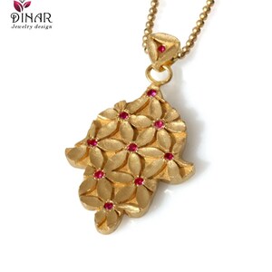 May include: A gold pendant necklace featuring a hamsa hand charm. The charm is adorned with small red gemstones and a floral pattern.