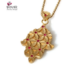 May include: A gold hamsa pendant necklace with a floral design and red gemstones. The necklace has a delicate chain.