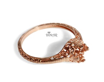 May include: A rose gold ring setting featuring an intricate filigree basket and a detailed engraved band. The metal surface displays a polished finish with ornate scrollwork patterns. The text DINAR is printed in black near the center.