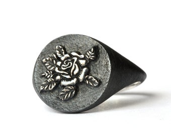 May include: A dark oxidized silver signet ring featuring a circular face with a raised, polished silver rose relief. The background of the ring face has a textured, matte finish, contrasting with the smooth, shiny petals of the flower.