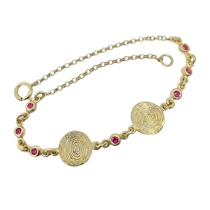 May include: A gold bracelet with three circular, textured pendants with a spiral design. The bracelet is decorated with small, round, red gemstones. The chain is delicate and has a lobster clasp closure.