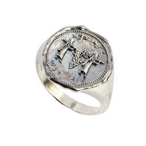 May include: A silver ring with a round, engraved design featuring two figures holding a bunch of grapes. The inscription around the design reads "על היין" in Hebrew.