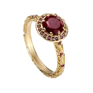 Può includere: Un anello d'oro con una grande gemma rotonda di colore rosso scuro al centro, circondata da pietre rosse più piccole. La fascia dell'anello è finemente dettagliata con un motivo floreale e ulteriori piccole gemme rosse.
