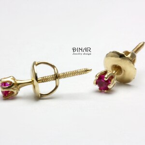 Może przedstawiać: Para złotych kolczyków sztyftów z jasnoczerwonymi kamieniami szlachetnymi. Kolczyki mają konstrukcję ze śrubą i są umieszczone na białym tle. Widoczna jest nazwa marki "DINAR jewelry design".