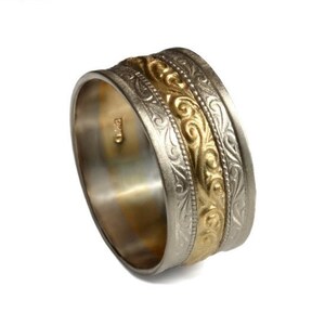 Op de afbeelding: Een zilveren en gouden trouwring met een ingewikkeld gegraveerd ontwerp. De ring is breed en heeft een verhoogde gouden band in het midden.