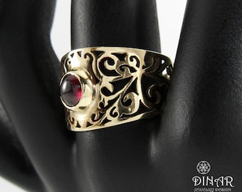 May include: A gold-toned ring featuring an intricate openwork filigree pattern and a single, round, deep red cabochon gemstone set in the center. The ring is displayed on a black mannequin hand, with the text DINAR JEWELRY DESIGN visible.