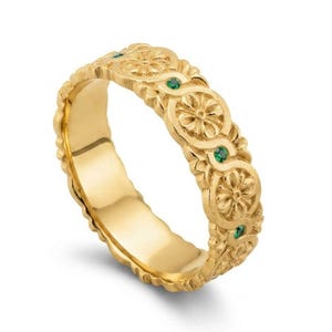 Pode incluir: Um anel de banda de ouro com um padrão floral em relevo repetido na superfície exterior. Pequenas pedras preciosas redondas de um verde vibrante estão cravadas entre cada motivo floral, e as bordas têm um acabamento texturizado.