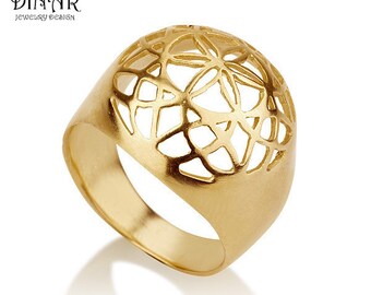 Puede incluir: Un anillo dorado con un diseño geométrico calado. El anillo presenta una banda ancha y una parte superior abovedada con un patrón repetitivo de formas interconectadas. El anillo está sobre un fondo blanco y la marca "DINAR JEWELRY DESIGN" es visible.