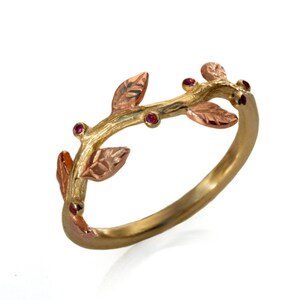 May include: A gold ring shaped like a textured branch with rose gold leaves and small, round, deep pink gemstones embedded along the band. The metal surface has a matte, organic finish that mimics the appearance of natural wood and foliage.