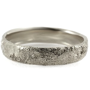 May include: A silver ring with a textured, organic surface. The ring has a band width of approximately 0.6 cm. The ring's surface has a rough, uneven texture, giving it a unique, handcrafted appearance. The ring is set against a white background.