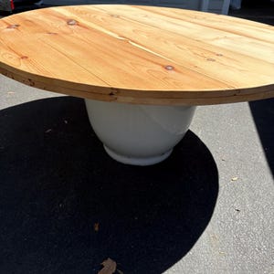 May include: A round wooden table with a light brown tabletop and a white base. The tabletop is made of planks of wood, and the base has a rounded shape. The table is outdoors on a paved surface.