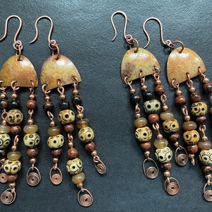 Puede incluir: Un par de aretes largos y colgantes con alambre de cobre y cuentas. Los aretes tienen una parte superior de cobre con un acabado martillado y tres hileras de cuentas. Las cuentas son de color marrón, negro y crema con un patrón de espiral.