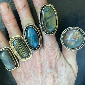 May include: Five rings with oval and round labradorite gemstones set in gold tone metal. The rings are worn on a hand.