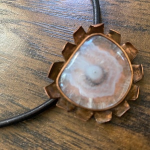 May include: A copper pendant with a white agate stone in the center. The pendant is attached to a black leather cord.