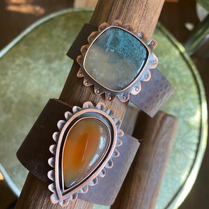 May include: Two handcrafted leather cuff bracelets. One features a square, light green and white stone set in a copper-colored, scalloped bezel. The other has a teardrop-shaped, orange stone in a similar setting. The leather is dark brown.