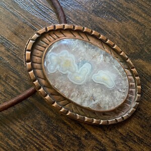 May include: A white agate stone with brown veins is set in a copper oval pendant with a textured border. The pendant is attached to a brown leather cord.