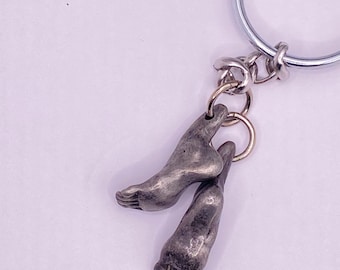 May include: Silver-toned keychain featuring two detailed foot charms. The charms are connected to a small ring, which is attached to a larger key ring. The feet are sculpted with realistic details, showcasing toes and arches.