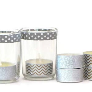 Może przedstawiać: Zestaw świeczek tealight z ozdobnymi papierowymi opakowaniami. Świece znajdują się w szklanych świecznikach z szarym i białym papierem w kropki wokół górnej części. Niektóre świece mają wzór chevron, a inne wzór kwiatowy w kolorze srebrnym.