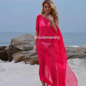 May include: A woman wearing a bright pink sheer beach cover-up over a white bikini top and a pink bikini bottom. The cover-up is flowing in the wind. The woman is standing on a sandy beach with large rocks in the background. The text "@solemareny" is visible on the cover-up.