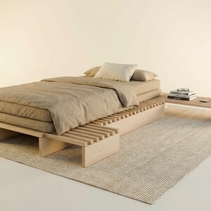 Könnte beinhalten: Ein niedriges Plattform-Bettgestell aus hellem Holz mit einem Lattenrost-Design, das sich zu einem Beistelltisch verlängert. Das Bett ist mit einer beigen Matratze, einer passenden Bettdecke und zwei Kissen bezogen, mit Büchern auf der Ablage.