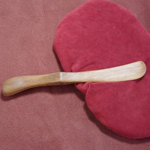 May include: A wooden butter knife with a curved handle, resting on a red fabric surface.